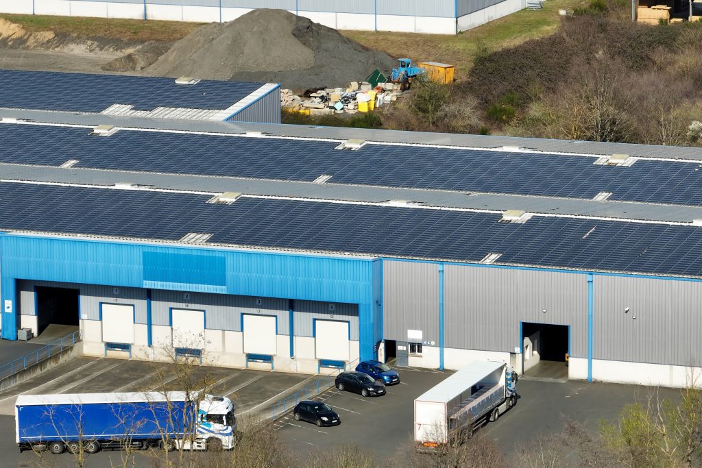 Two delivery lorries entering a blue and grey warehouse with three rows of solar panels on the roof.