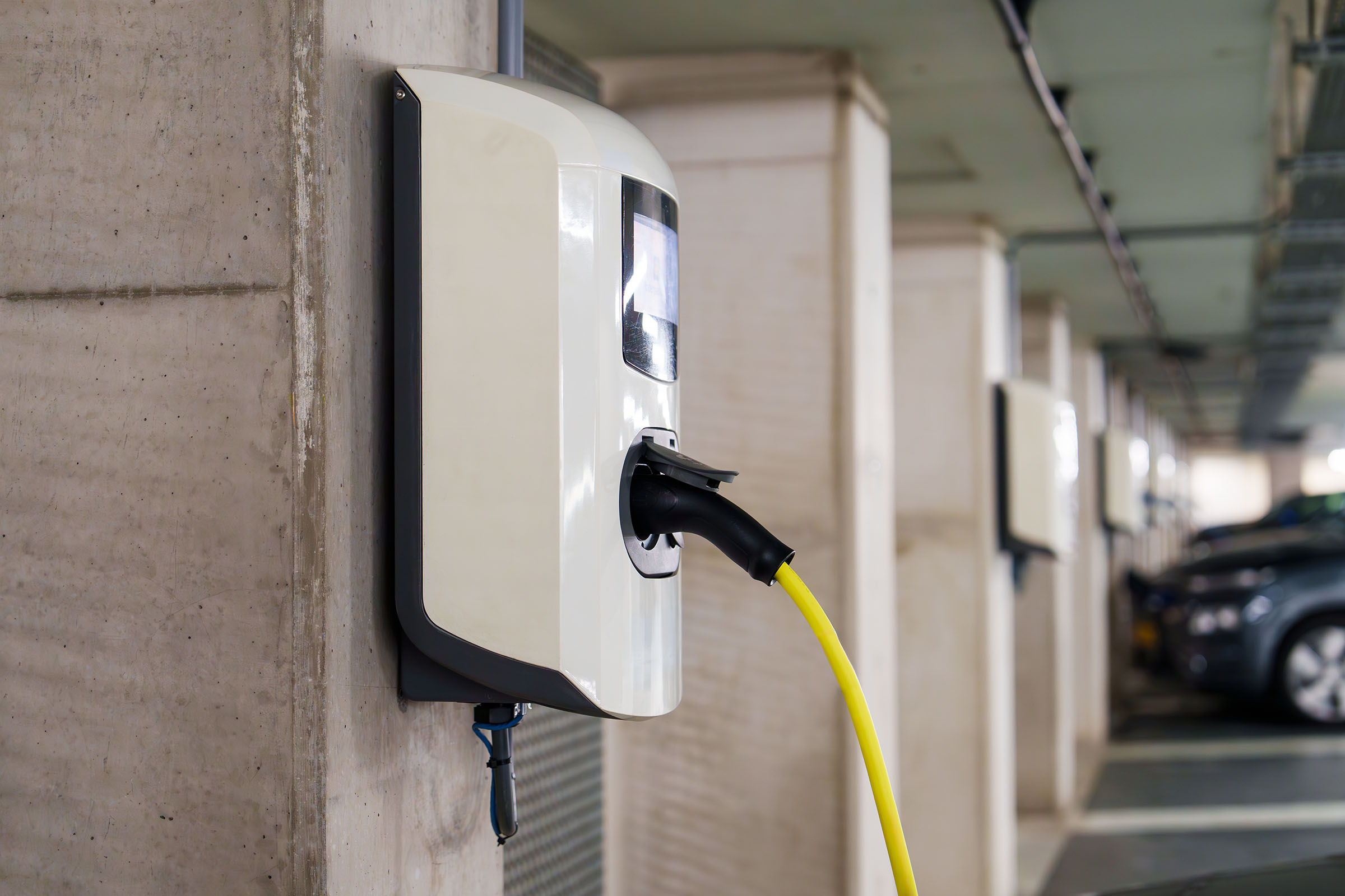 A single electric vehicle charging point mounted on a concrete column in a car park, in use and connected to a yellow charging cable.