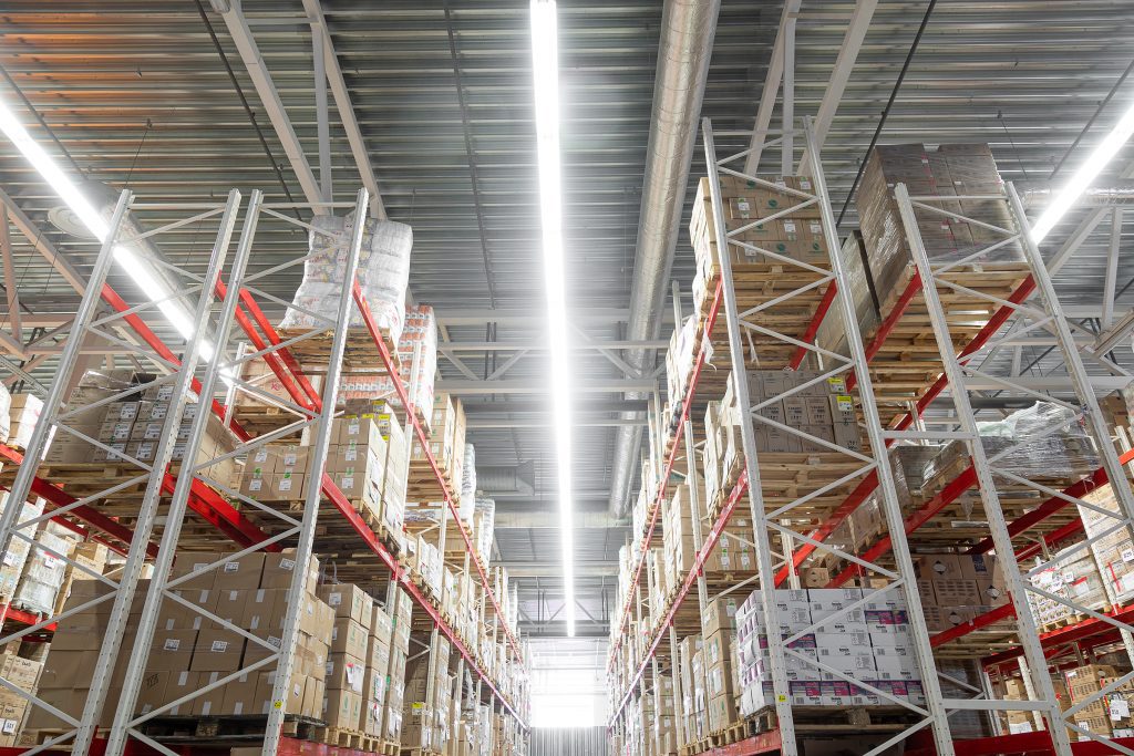 Interior of a brightly-lit warehouse, with tall racking on either side, stocked with cardboard boxes.